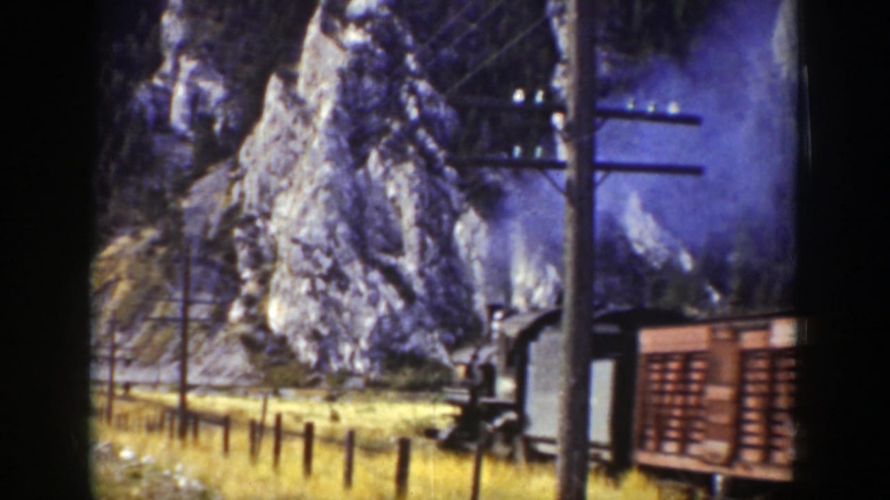 1939 Wyoming 8mm frame showing a steam train beside rocky cliffs, yellow grass, utility poles, and overhead wires.
