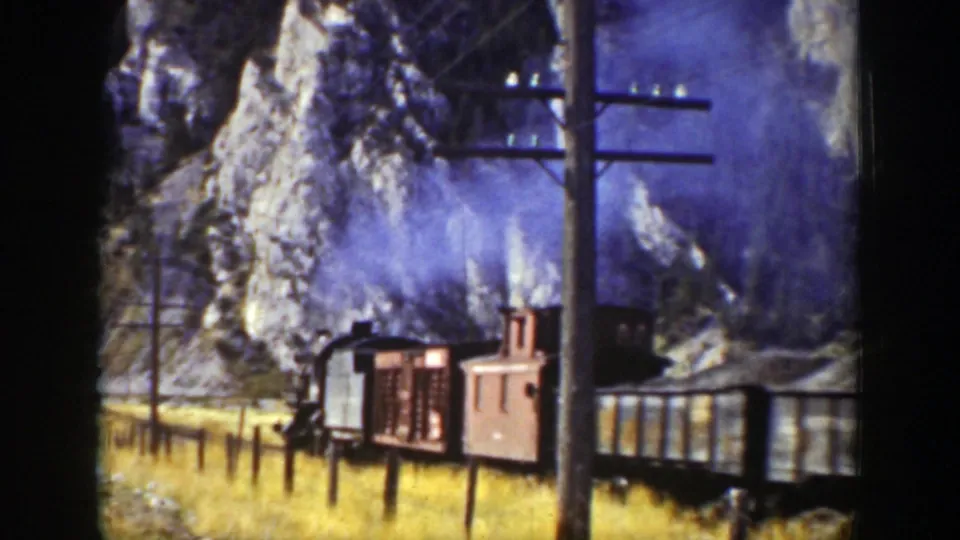 1939 Wyoming 8mm frame with a locomotive, freight cars, utility poles, wires, grass, and steep rocky terrain.