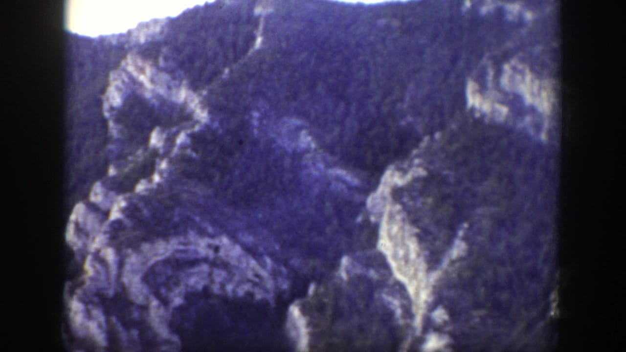 Rocky mountain slope with dark trees and pale cliff faces seen through a film gate.