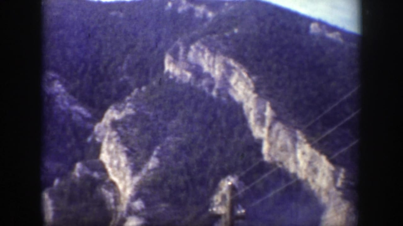 Mountain cliffs and wooded slopes with thin diagonal wires crossing the right side.