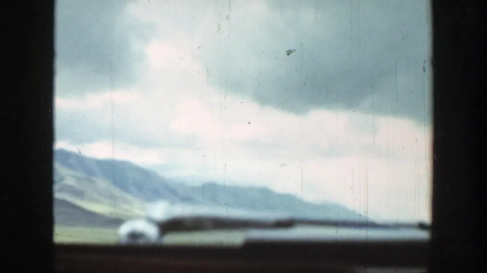 1939 Wyoming travel footage frame with a blurred car-window edge, open field, distant ridge, and overcast sky.