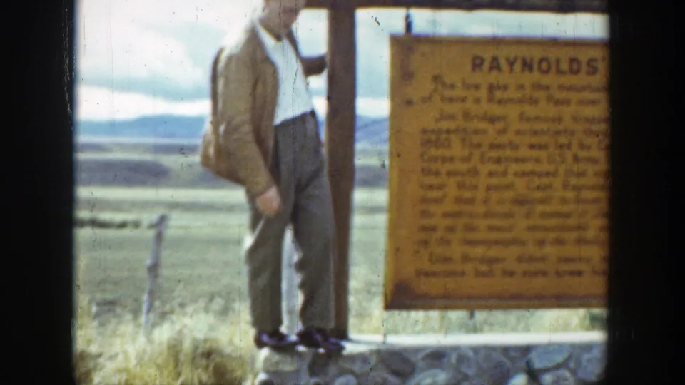 1939 Wyoming frame of a man standing near the Reynolds Pass marker, with grassland, fence posts, and distant hills behind him.