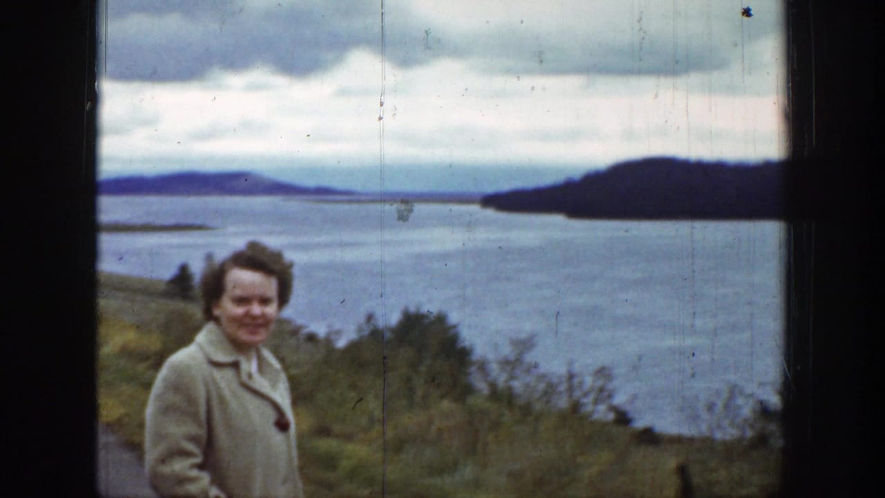 In 1939 8mm home-movie footage, a woman in a tan coat stands by a roadside water overlook and smiles toward the camera.