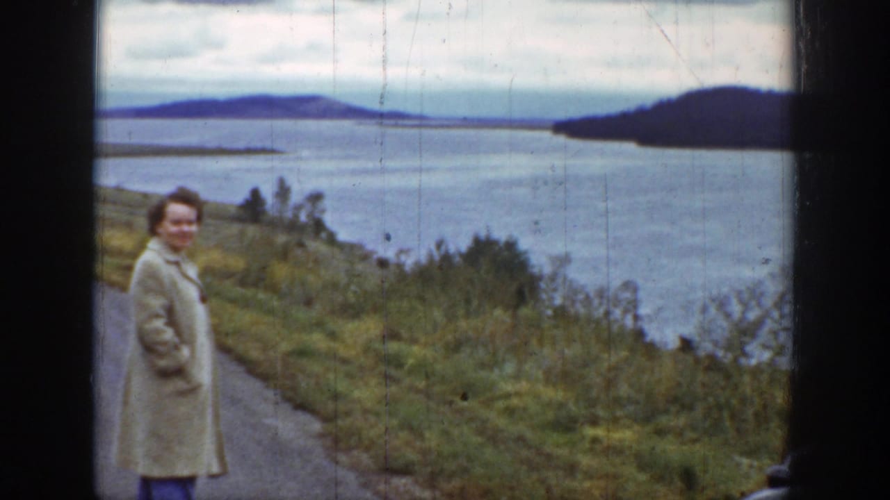 Woman in a light coat stands beside a road overlooking a lake and distant hills.