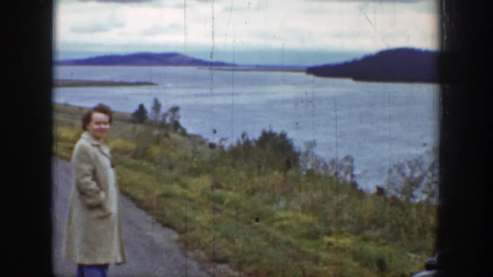 Adult woman in a light coat stands near a roadside above a Wyoming lake in 1939, with cloudy sky and distant hills behind her.