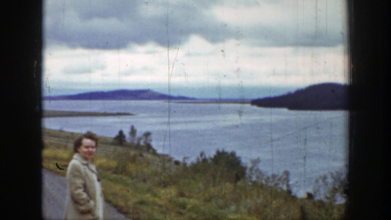 Woman stands near the roadside with a broad lake, grassy bank, and dark hills beyond.