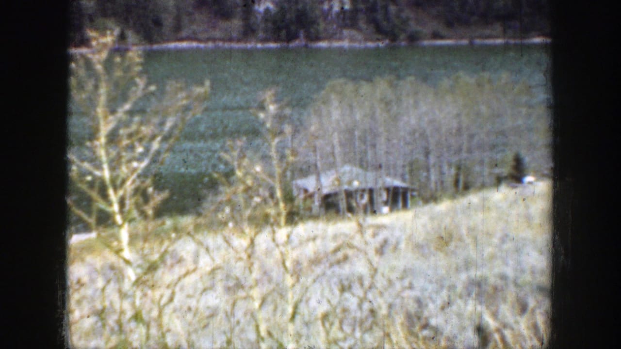 1939 Wyoming home-movie frame showing a small cabin among leafless trees, with water and distant terrain beyond.