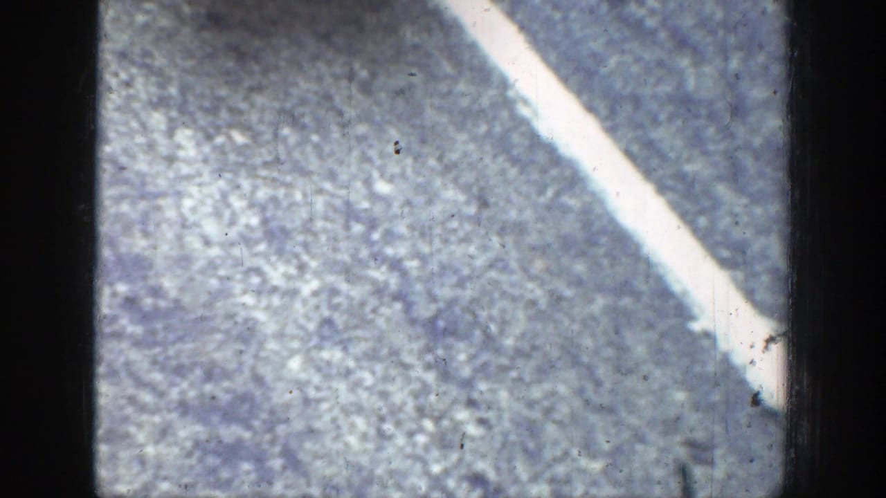 Blurred aerial view of pale ground or hillside with a bright diagonal line and film grain.