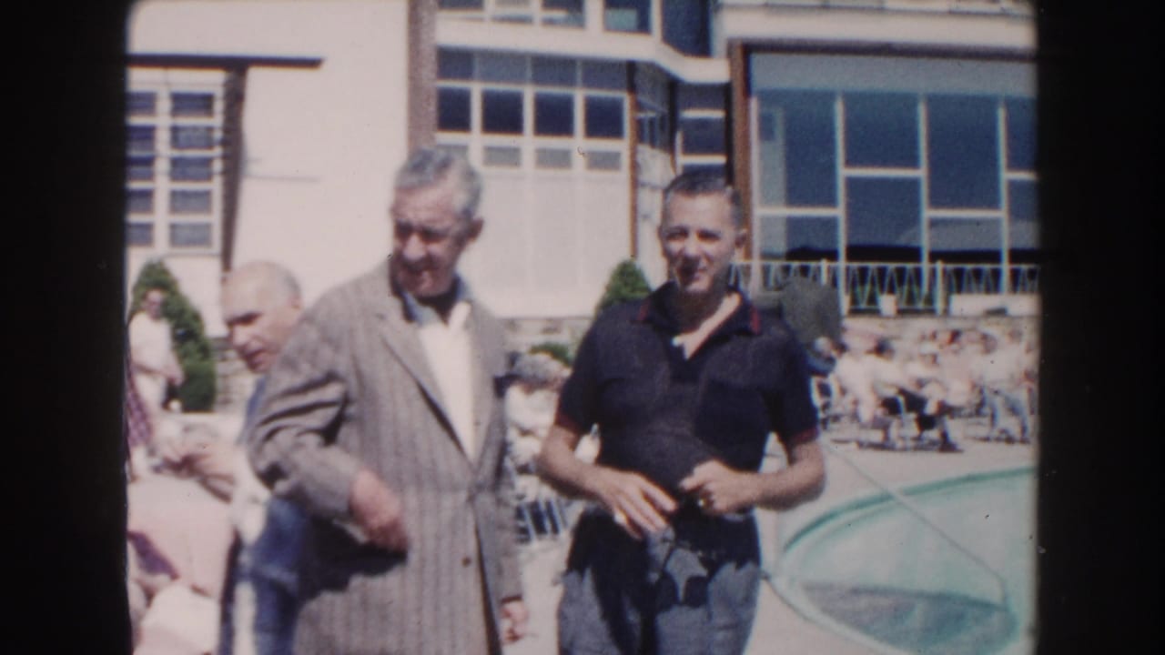 1960 Miami home-movie frame of elderly men gathering outdoors near a swimming pool and large-windowed building.