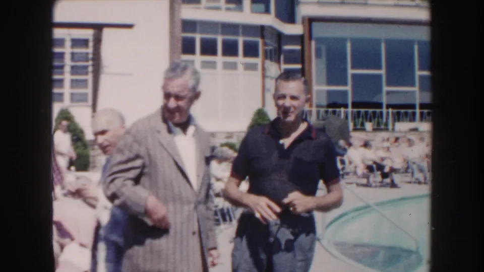 1960 Miami home-movie frame of elderly men gathering outdoors near a swimming pool and large-windowed building.