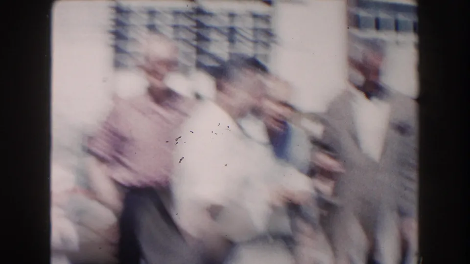 Soft, motion-blurred 1960 Miami home-movie frame of men shifting position near a poolside building.
