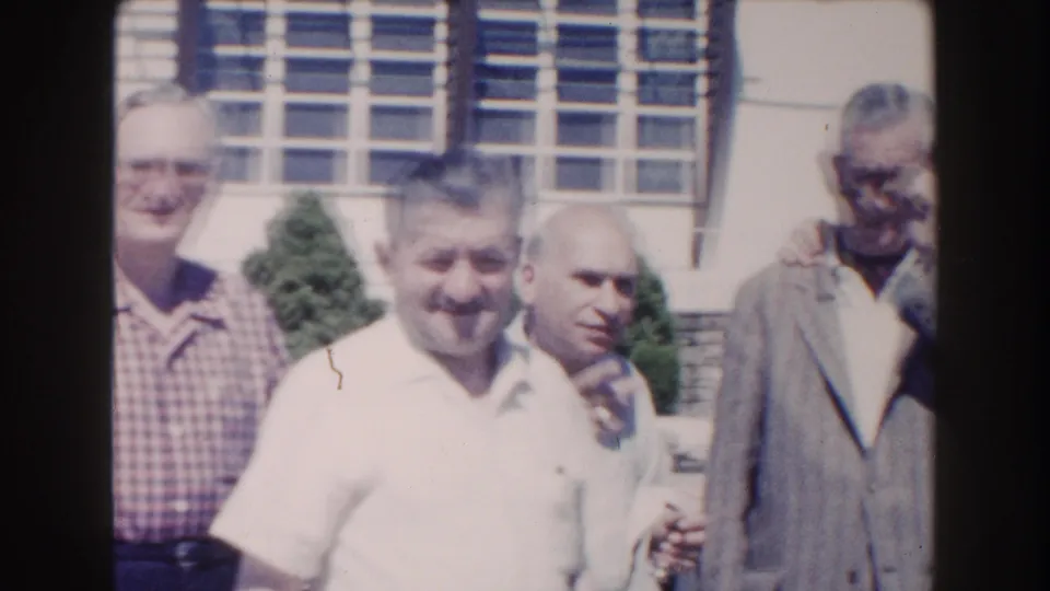 1960 Miami frame of older men standing close together outdoors, with windows and poolside landscaping behind them.
