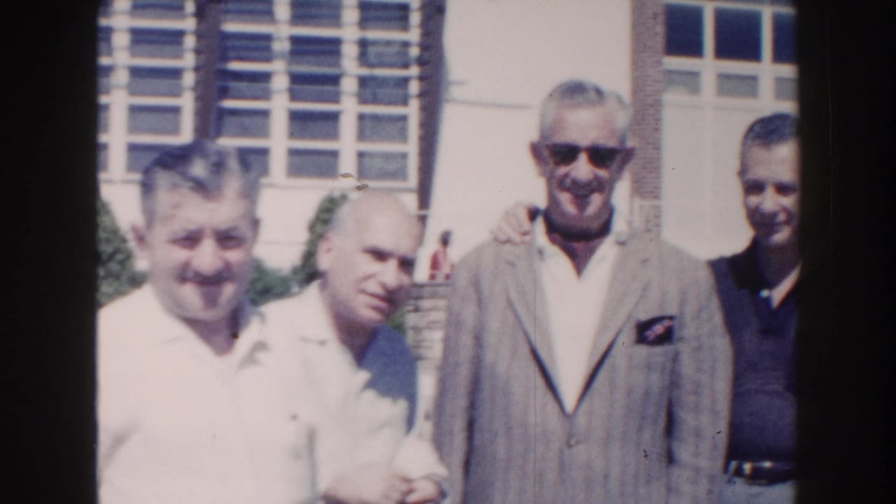 1960 Miami home-movie frame of adult men standing in bright sun near large windows, including a man in a gray jacket and sunglasses.