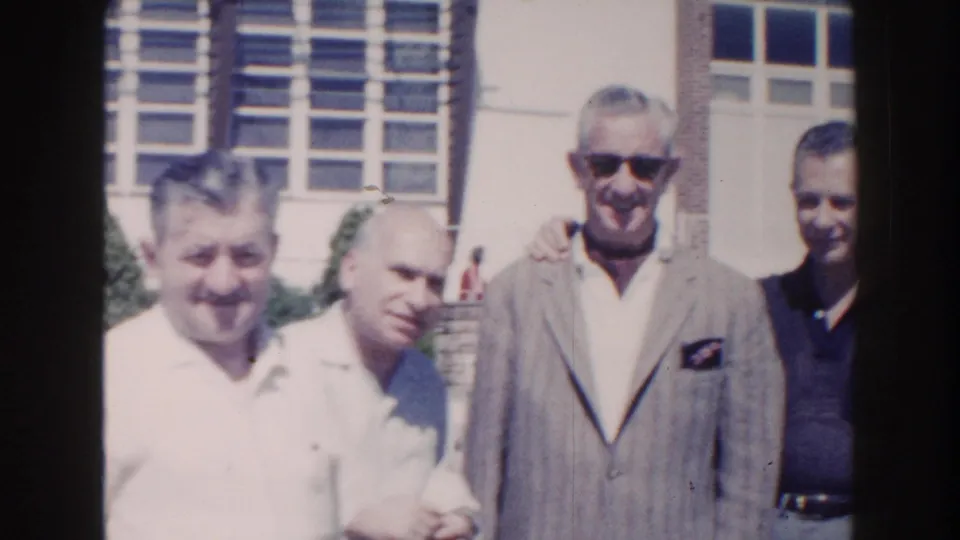 1960 Miami home-movie frame of adult men standing in bright sun near large windows, including a man in a gray jacket and sunglasses.