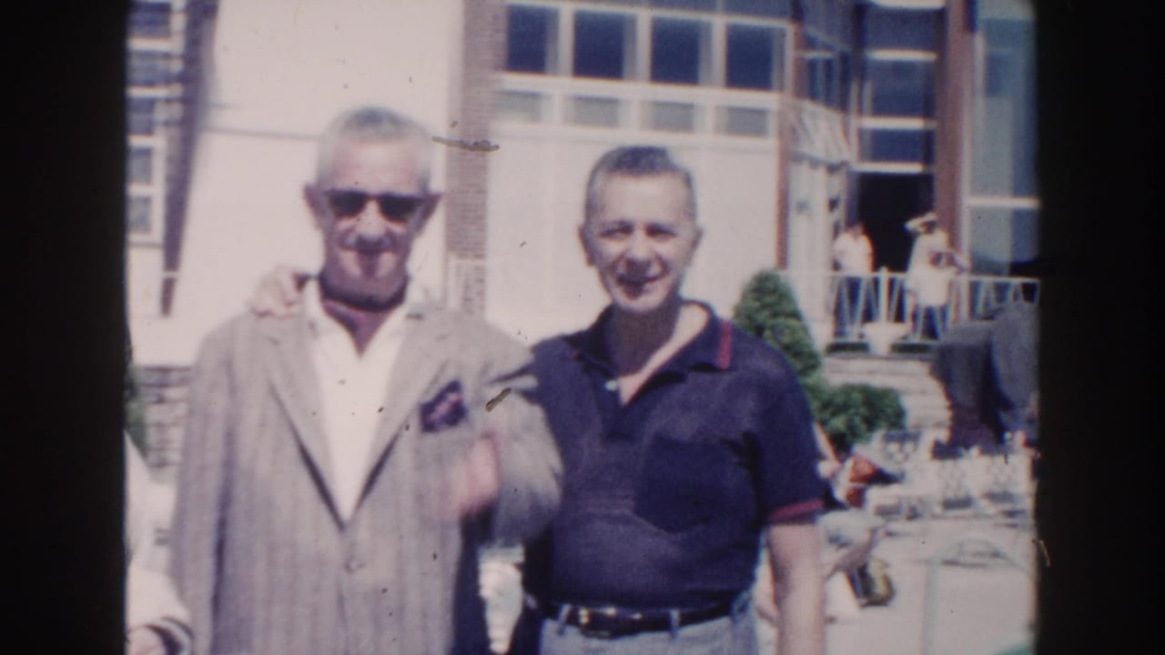 Two men stand shoulder to shoulder outdoors, one wearing sunglasses and a gray jacket.