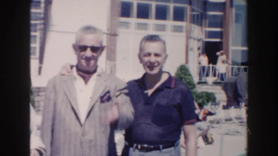 Two adult men face the camera in a sunny 1960 Miami home-movie frame, with a building entrance and windows behind them.