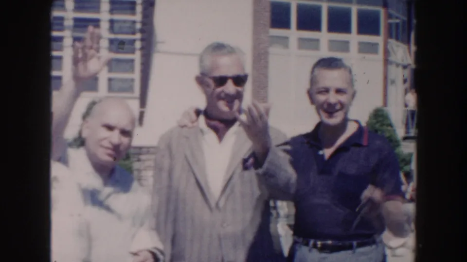 1960 Miami home-movie frame showing adult men smiling and waving in bright daylight outside a building with large windows.