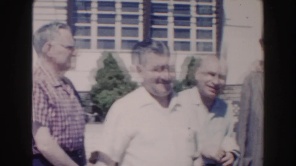 Adult men pass through the frame in a sunny 1960 Miami home-movie shot, with shrubs and building windows in the background.