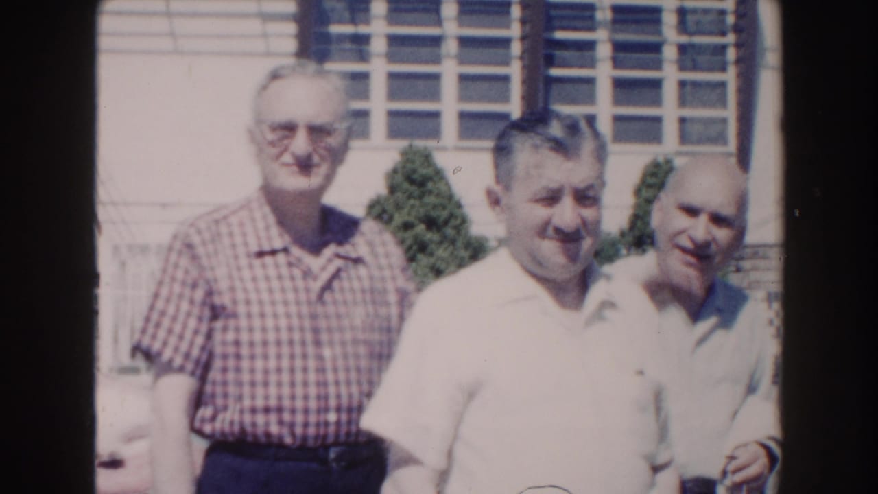 Three older men in casual 1960s clothing stand together outdoors in Miami, with louvered windows behind them.
