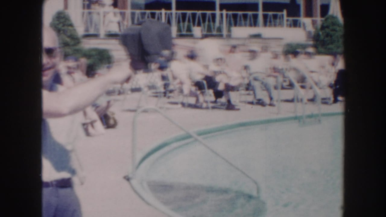 Man in sunglasses points near a blue swimming pool with seated people in the background.