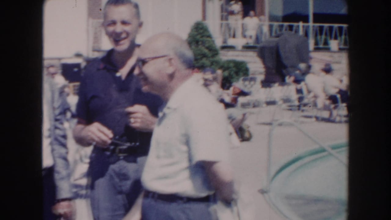 Two adult men stand beside a swimming pool while one smiles toward the camera.