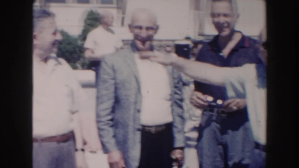 1960 Miami home-movie frame showing three adult men by the pool while an offscreen person points across the scene.