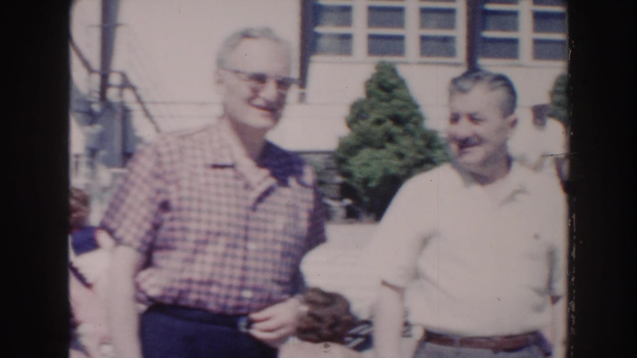 Two older men stand outdoors near a sunlit building and shrubbery.