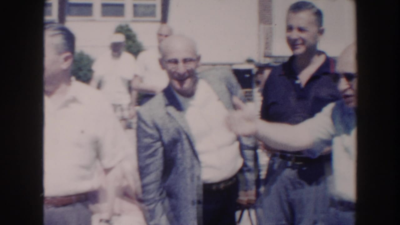 Older men gather outdoors in 1960 Miami, smiling and gesturing near a building with large windows.