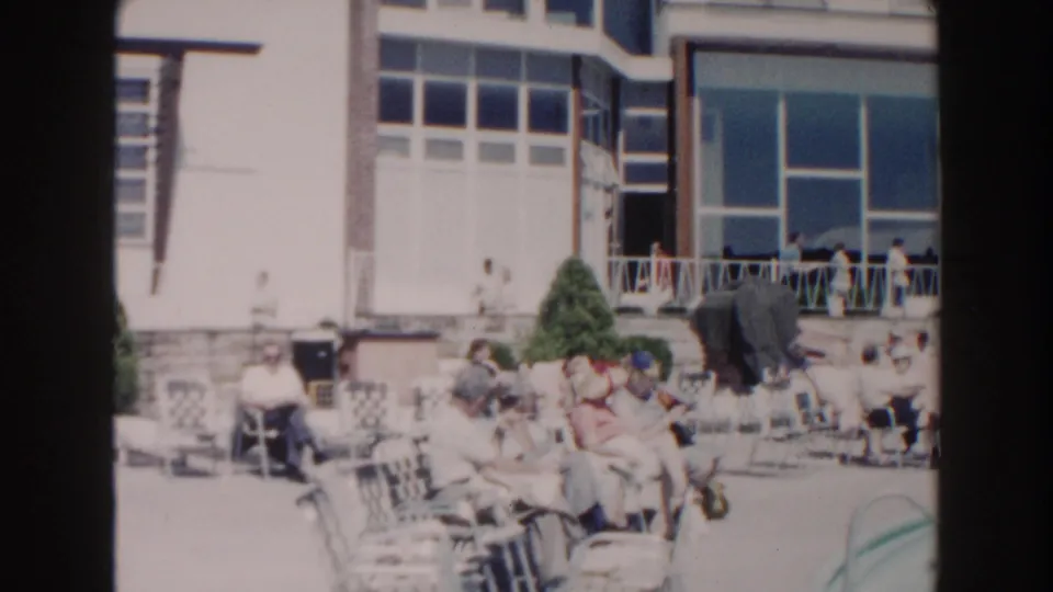 A poolside patio in 1960 Miami shows seated adults, lounge chairs, railings, shrubs, and a sunlit residential building.