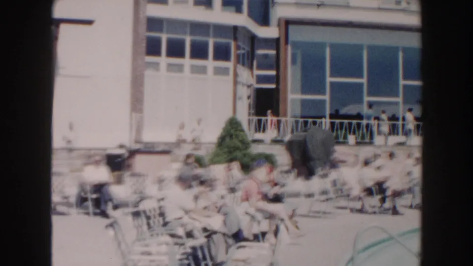 The Miami poolside area shows adults seated near lounge chairs, a manicured shrub, railings, and large building windows.