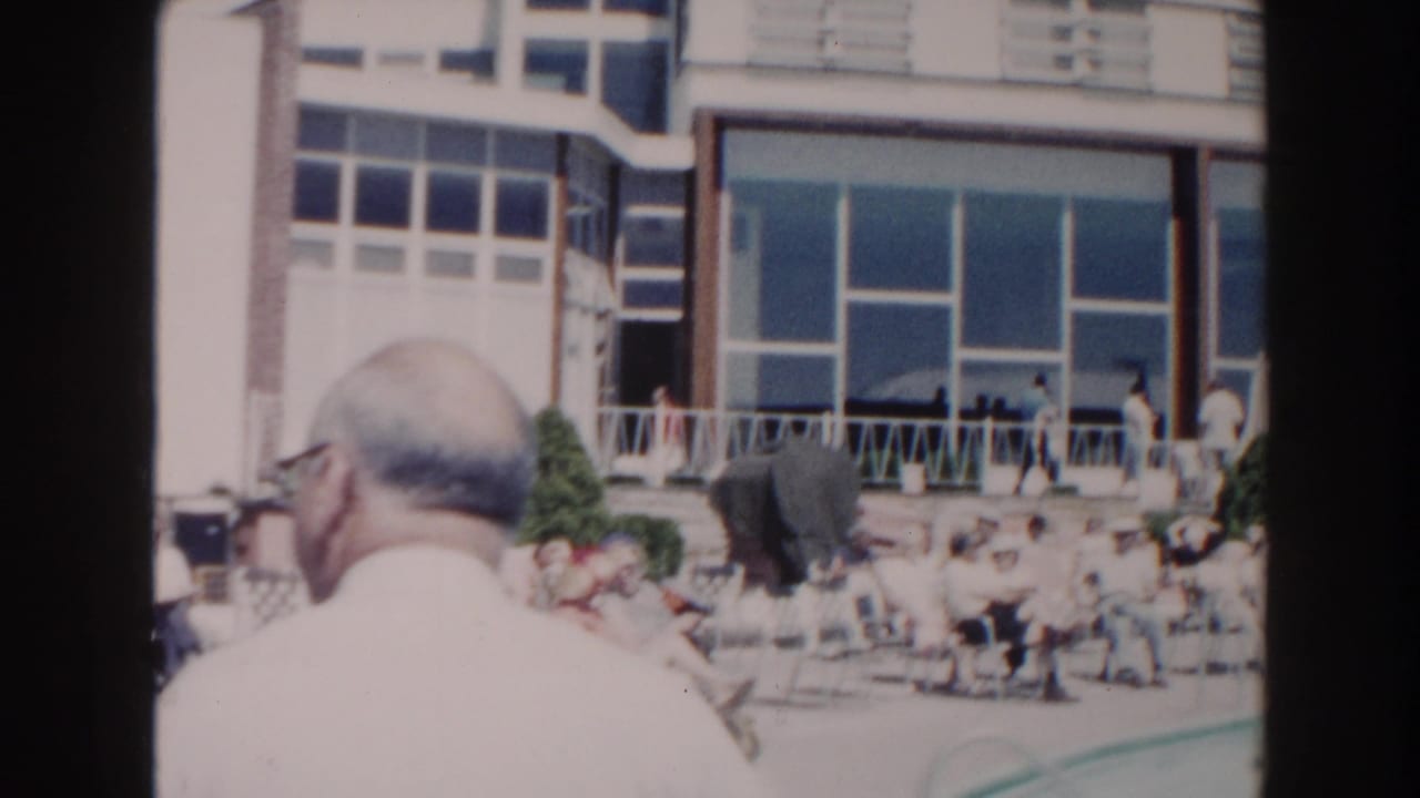 Guests lounge beside a pool in front of a glass-fronted building, with a man seen from behind in the foreground.