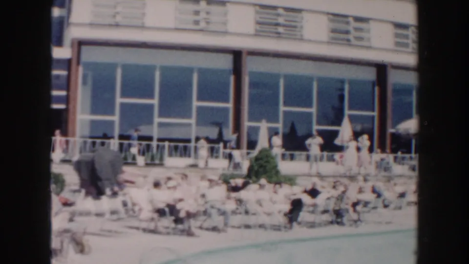 1960 Miami poolside view showing sunbathers, chairs, umbrellas, railing, and a modern glass-fronted resort building.