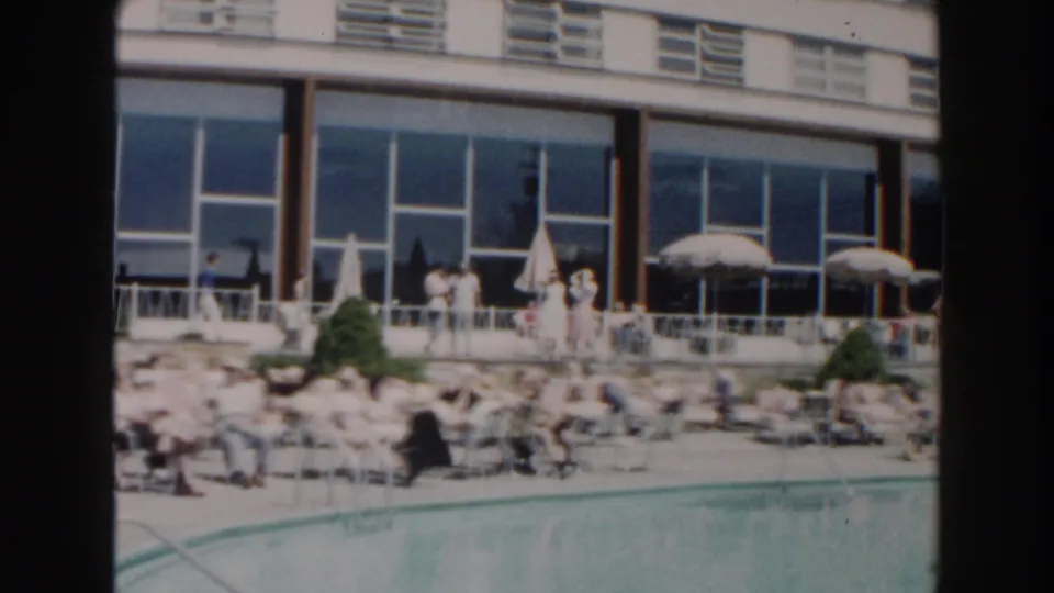 1960 Miami home-movie frame with resort guests lounging beside the pool near umbrellas, shrubs, railings, and large windows.