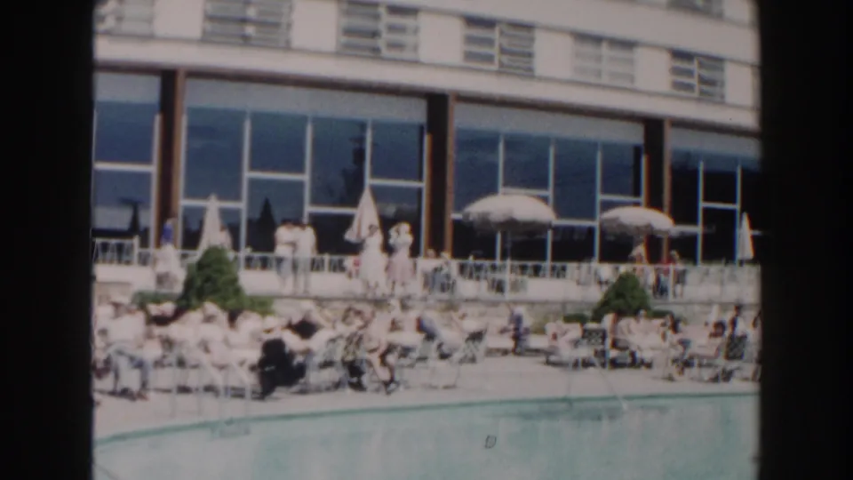 1960 Miami hotel poolside with sunbathers, white umbrellas, patio seating, and a modern glass-fronted building.