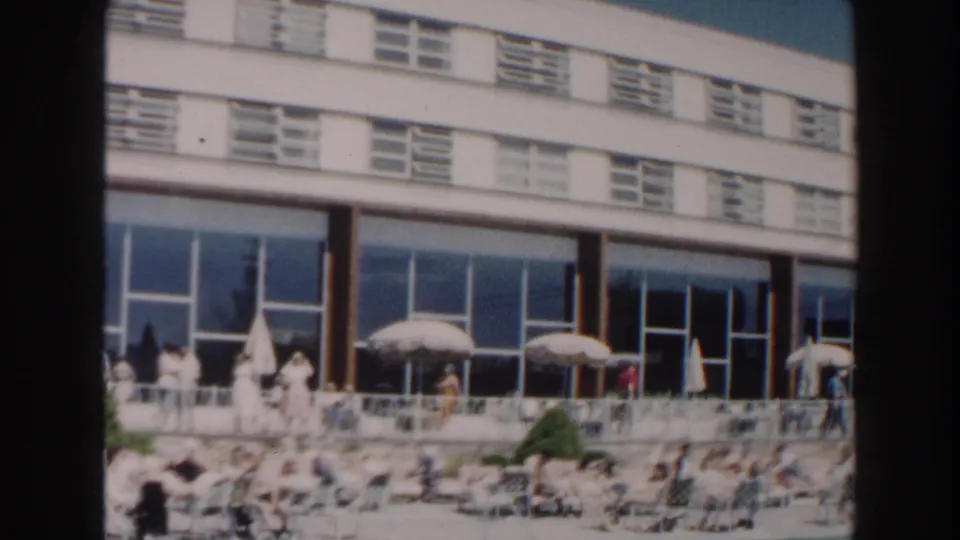 1960 Miami resort pool area with guests seated near umbrellas under a modern hotel facade.