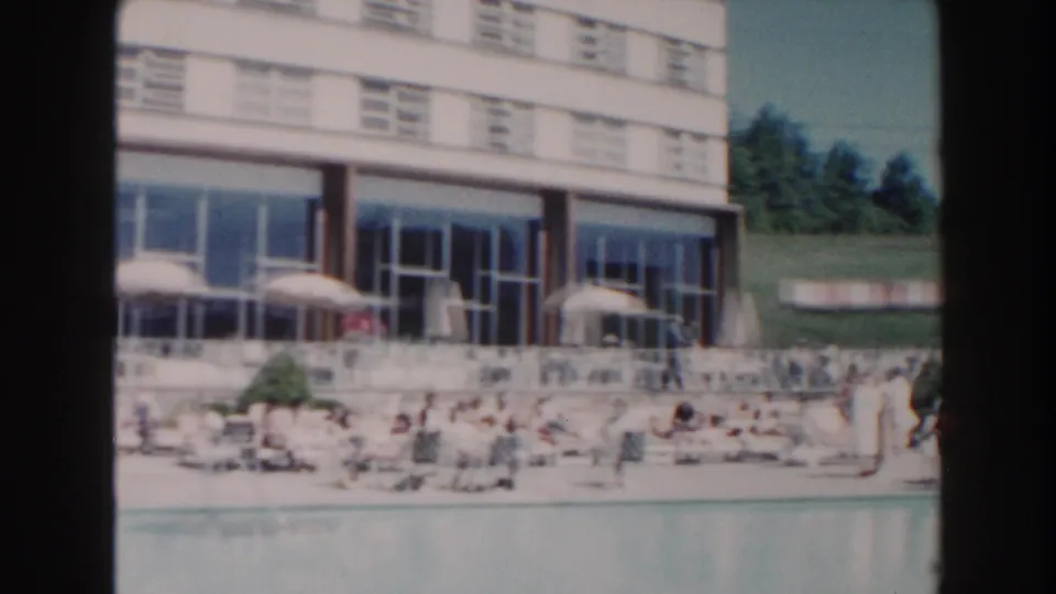 1960 Miami hotel pool scene with soft 8mm color, sunbathers, umbrellas, and a glass-fronted patio.