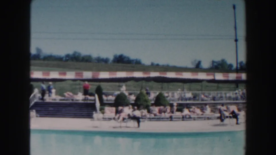 1960 Miami public pool view with visitors on lounge chairs, a striped awning, steps, a tall pole, and open grass behind the deck.