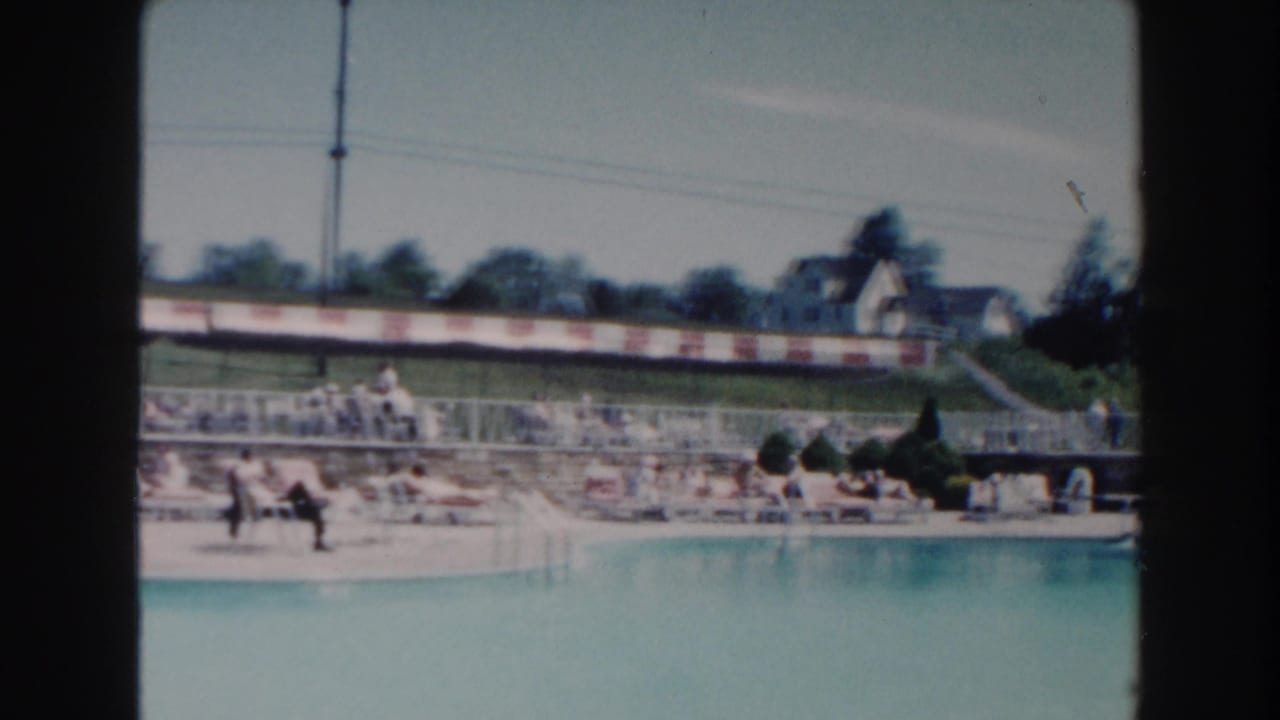 Poolside lounge area with seated visitors, striped barrier, trees, houses, and bright sky.
