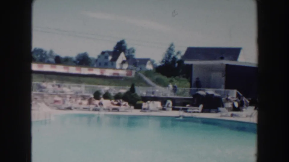 1960 Miami poolside view showing pale blue water, lounge chairs, distant visitors, a grassy slope, houses, trees, and a low building.