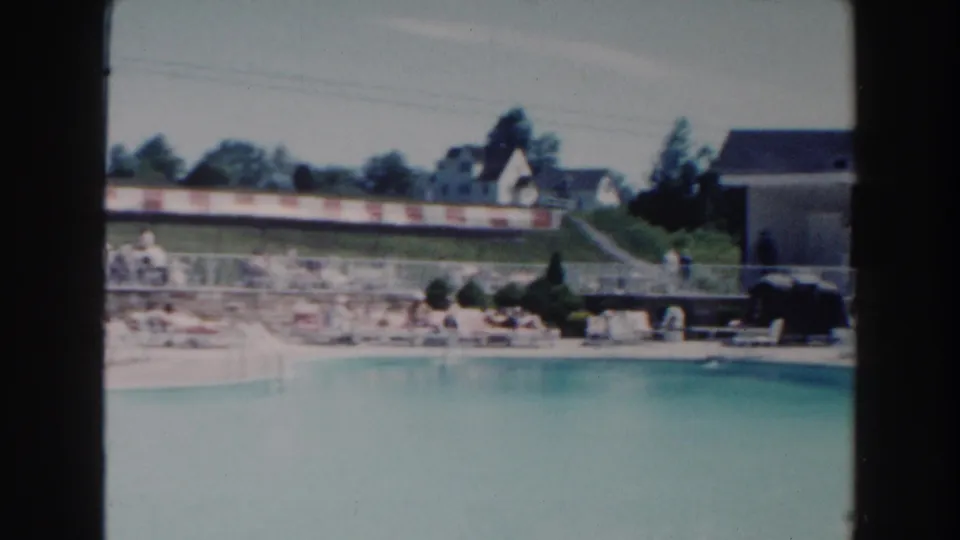 1960 Miami pool scene with blue water in the foreground, people near lounge chairs, a striped barrier, trees, houses, and a low building.