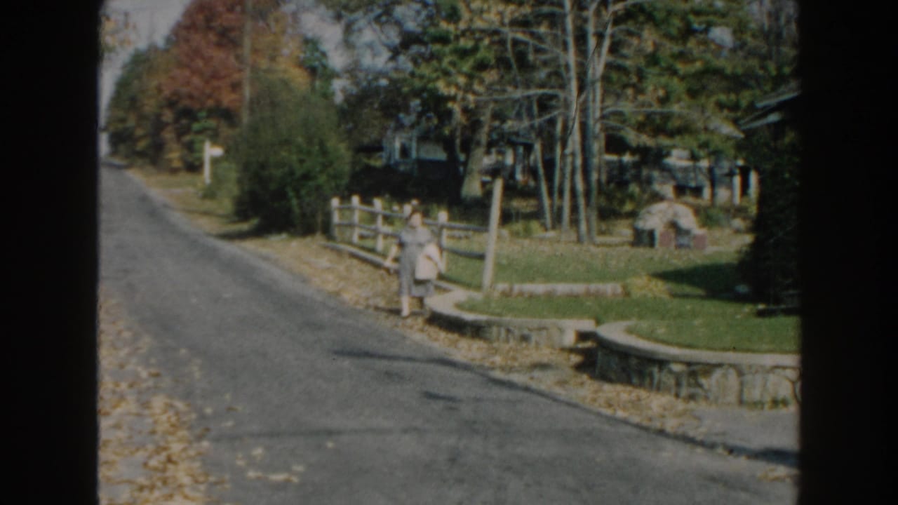 1960 Miami home-movie frame showing a woman in a gray dress walking along a sunny residential street.