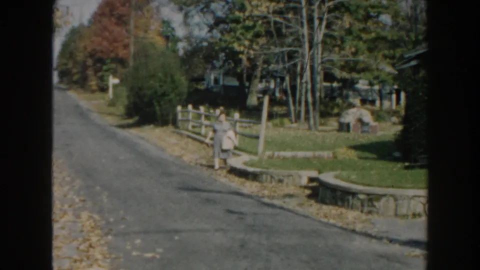 1960 Miami home-movie frame showing a woman in a gray dress walking along a sunny residential street.