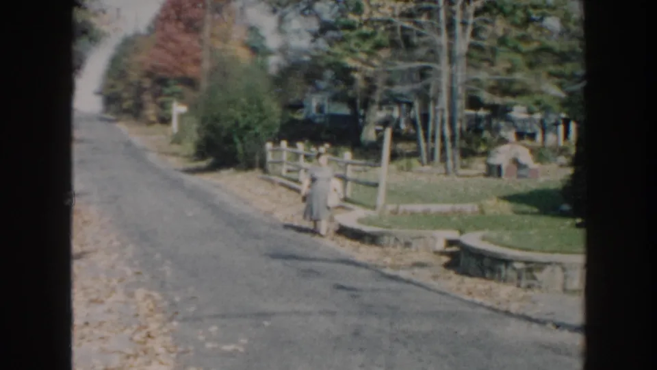 1960 Miami frame of a woman in a gray dress near a wooden fence and landscaped yard beside a quiet road.