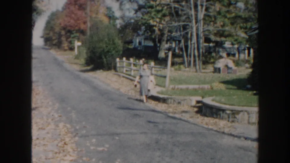 1960 Miami home-movie frame showing the woman walking closer along a residential street with autumn leaves at the curb.