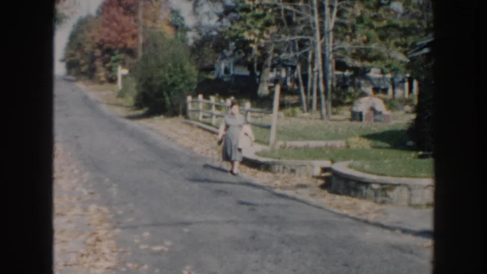 1960 Miami frame showing a woman walking near a low fence and tree-lined yard on a sunny suburban street.
