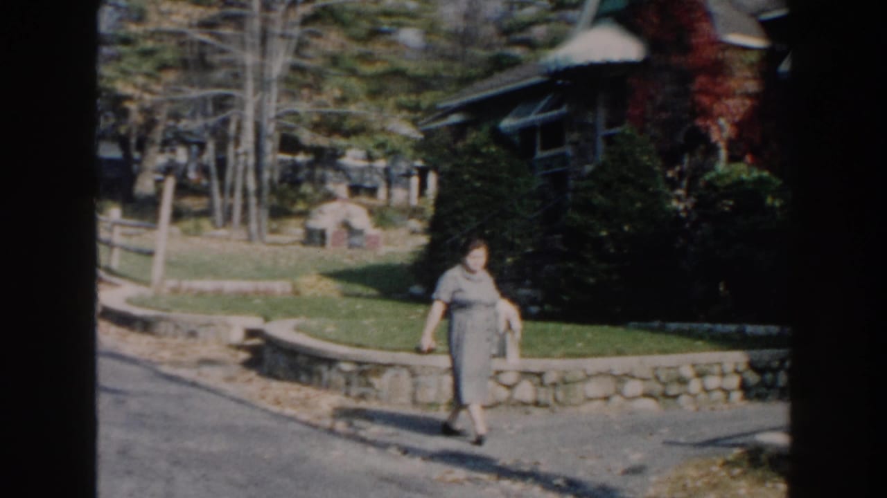 In 1960 home-movie footage identified as Miami, a woman in a gray dress walks past a stone ledge, lawn, trees, and house exterior.