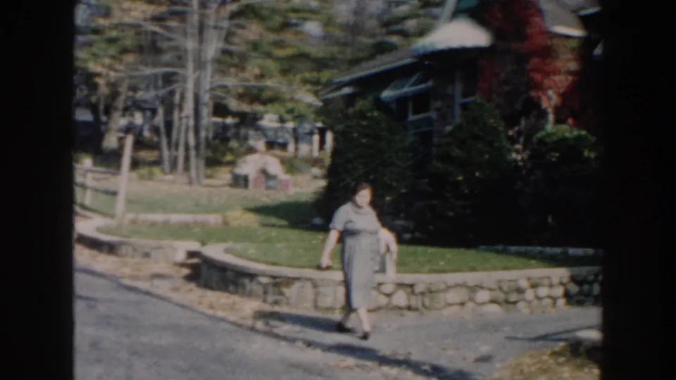 In 1960 home-movie footage identified as Miami, a woman in a gray dress walks past a stone ledge, lawn, trees, and house exterior.