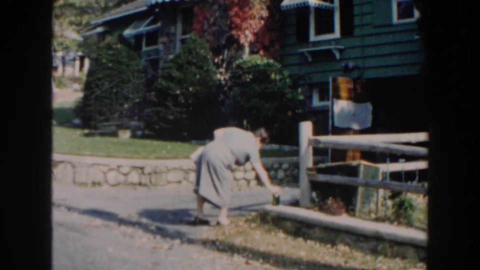 The woman bends toward the roadside ledge, appearing to place a green bottle near the stonework and wooden fence.