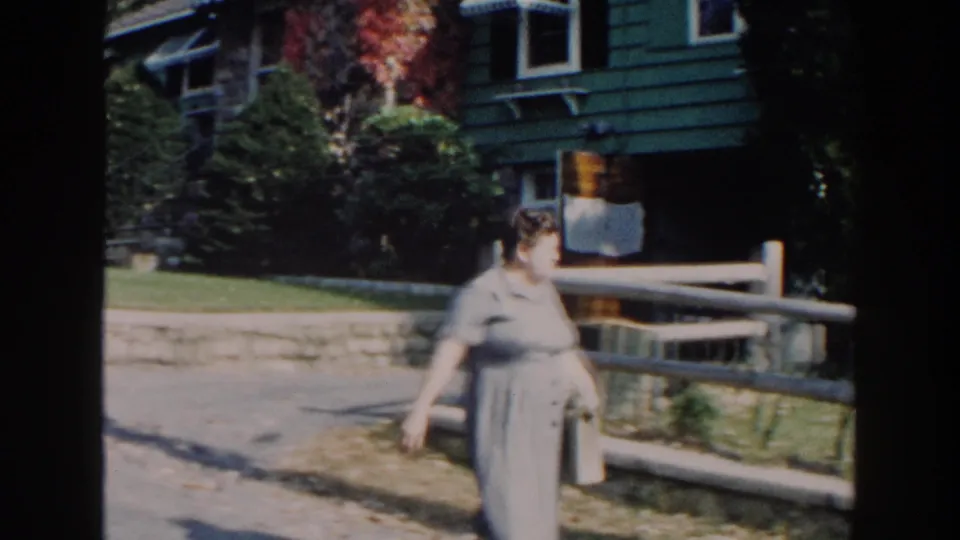 After setting down the bottle, the woman walks past the wooden fence with the house exterior and stone ledge still visible.