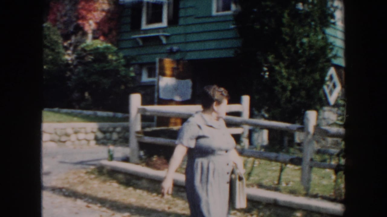 Woman in a light dress walks beside a wooden fence near a green-sided house.